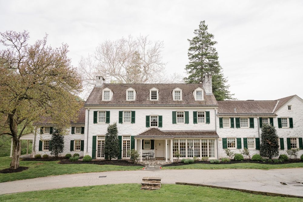 Elegant white colonial mansion with black shutters, manicured grounds, and tall evergreen tree on overcast day.