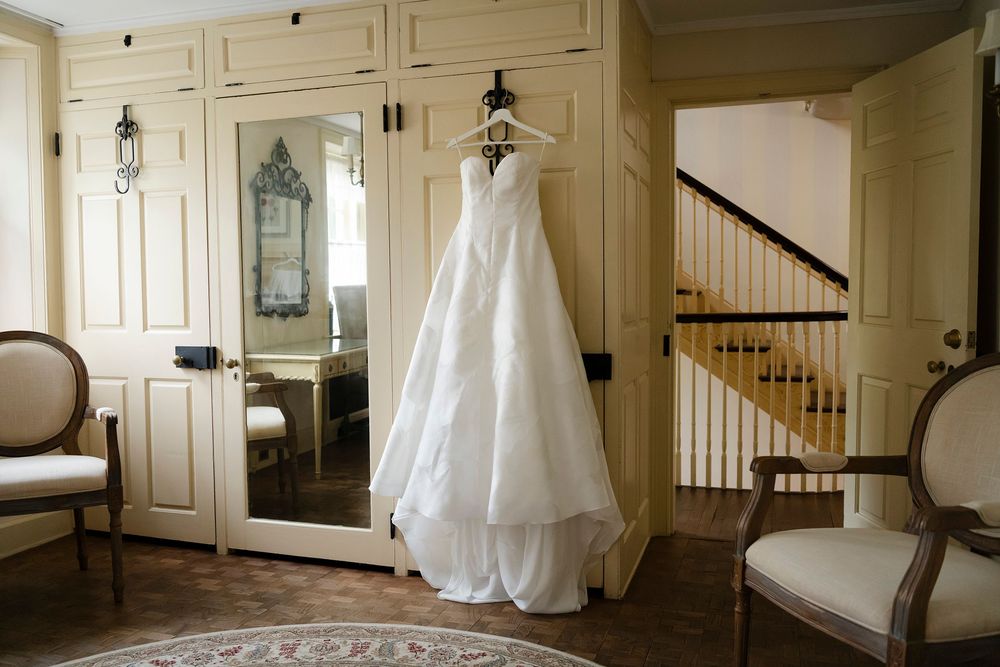 White wedding dress hanging on a hanger in an elegant room with mirrored wardrobe and wooden staircase.