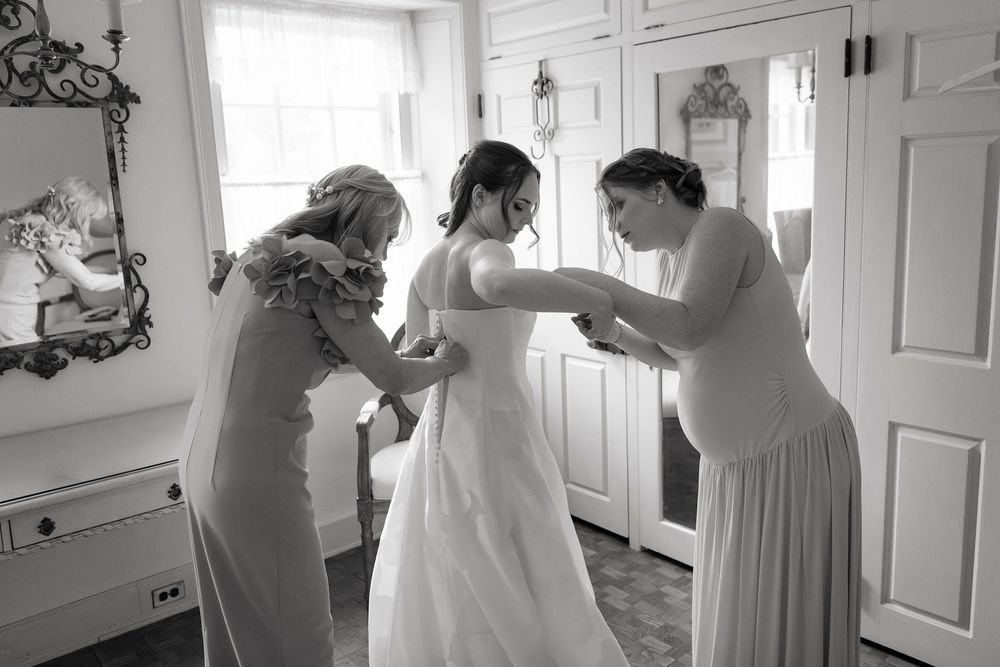 Black and white photo of a bride being helped into her wedding dress by two women in a bright, elegant room.
