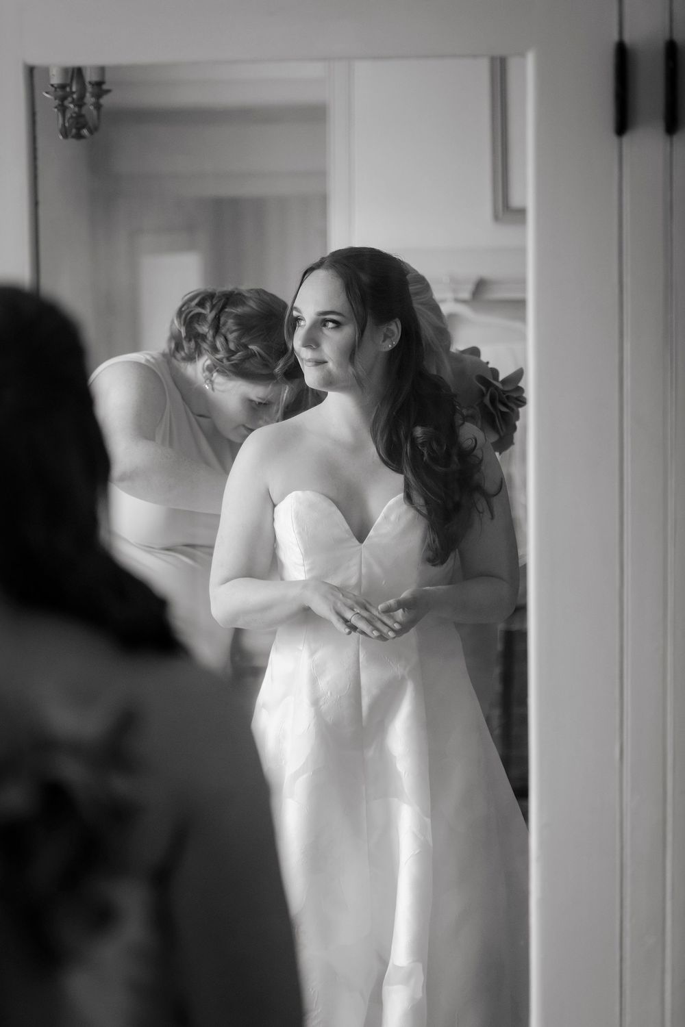 Black and white photo of a bride in a strapless gown being helped by a bridesmaid, gazing thoughtfully in a mirror.