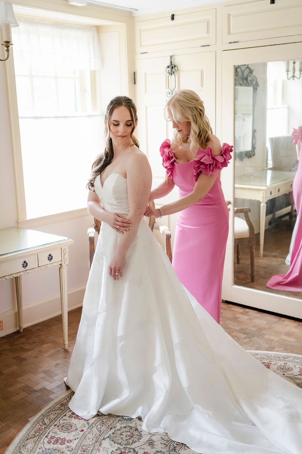 Bride in white gown being helped by bridesmaid in pink ruffle dress in elegant bridal suite.