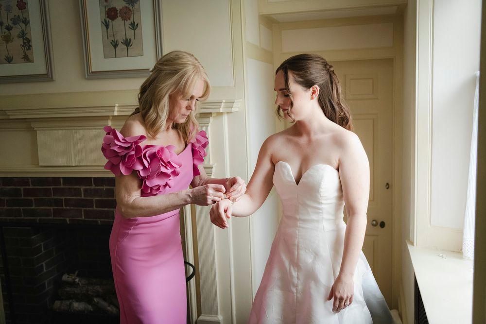 Bride in strapless white gown receives jewelry help from woman in pink ruffled dress in elegant room.