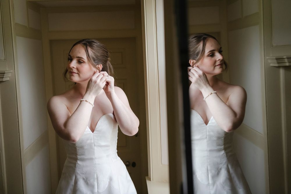 Bride in strapless lace wedding dress putting on earrings reflected in mirror while getting ready.
