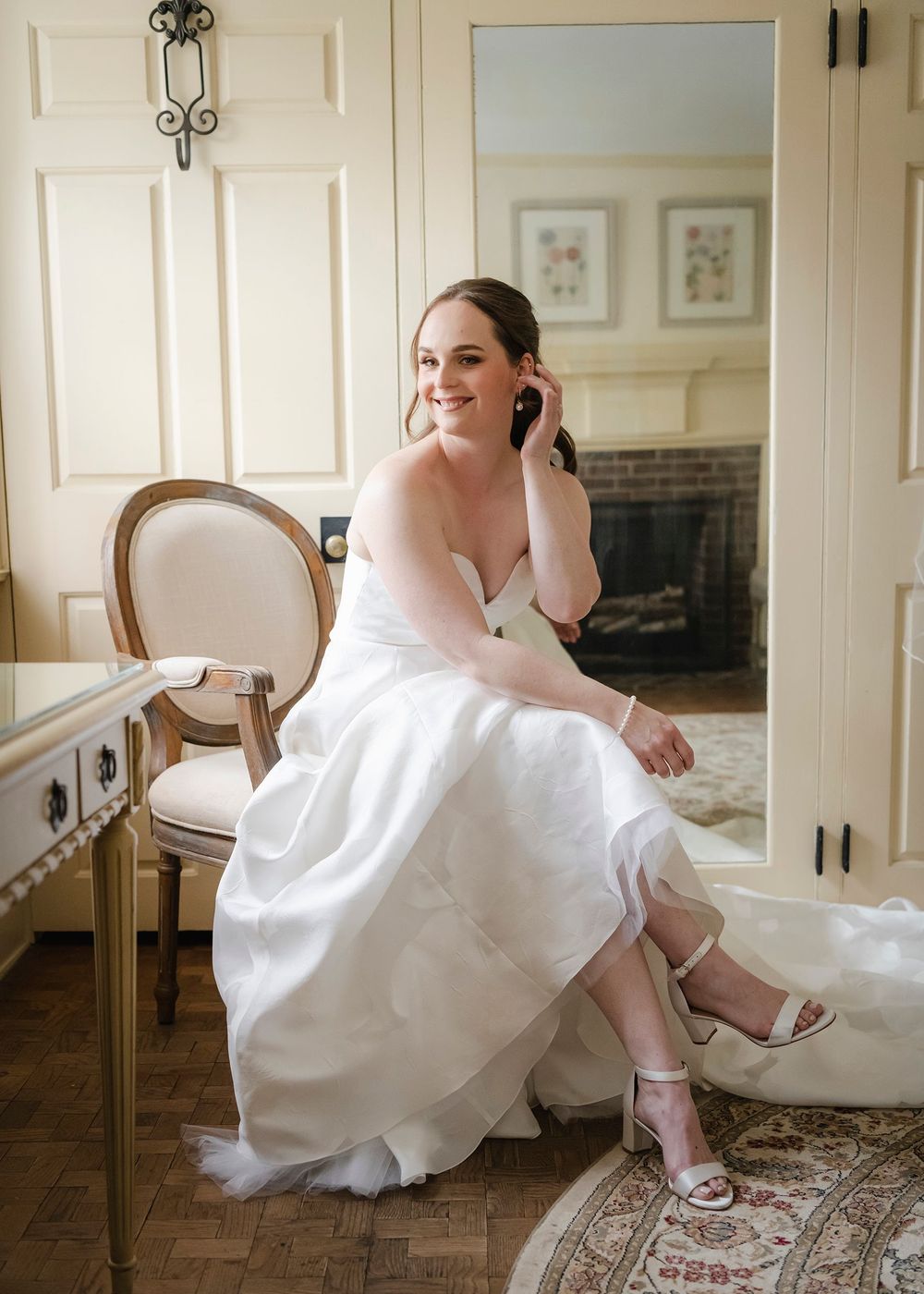 Bride in white gown sitting at vanity, touching earring, smiling, with elegant room and fireplace reflected in mirror.