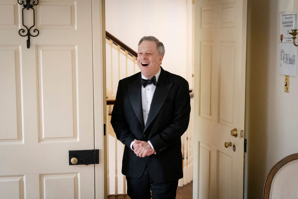Elderly man in black tuxedo and bow tie laughing joyfully while standing in a doorway at a formal event.