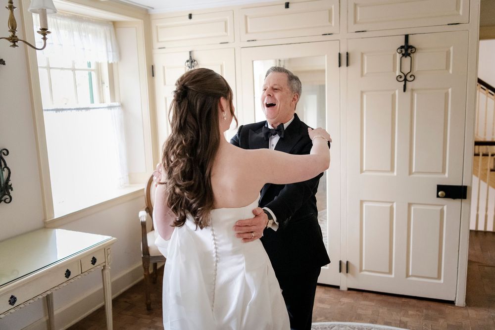 Father reacts with joy seeing bride in white wedding gown during emotional first look moment indoors.