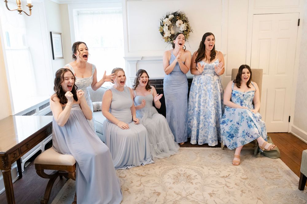 Bridesmaids in blue gowns laughing and celebrating together in an elegant white room during wedding preparations.
