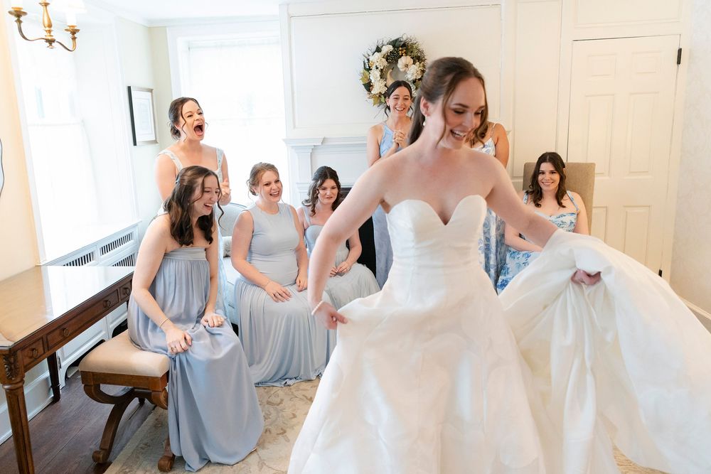 Bride in white ball gown twirls as bridesmaids in silver dresses react with joy in elegant wedding preparation room.