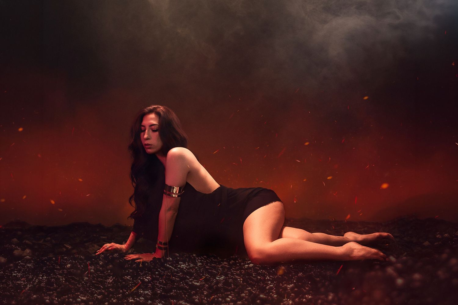 Woman in black dress poses dramatically on dark ground surrounded by red flames and smoke.