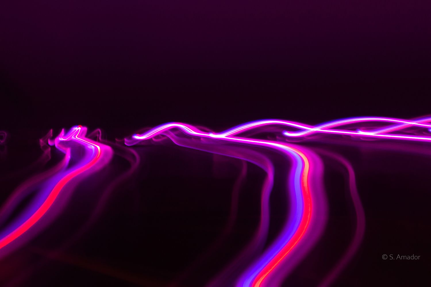 Abstract long exposure photography of vibrant pink, purple, and red light trails streaking across a dark background.