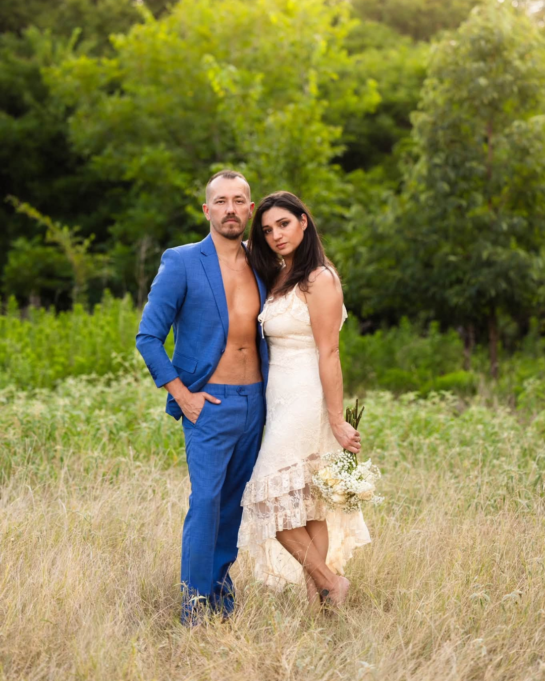 A couple in wedding attire embraces outdoors, the groom in an open blue suit and bride in a white lace dress holding flowers.