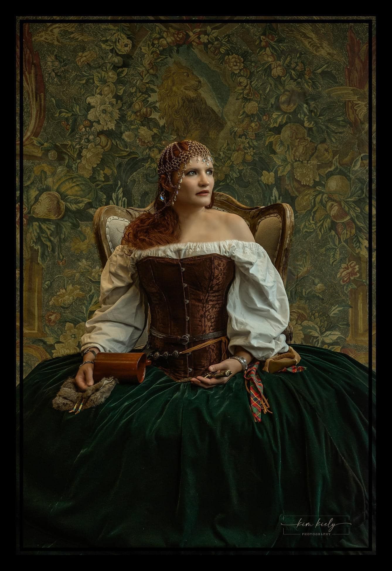 Woman in Renaissance corset gown and jeweled headpiece seated in ornate chair before floral tapestry backdrop.