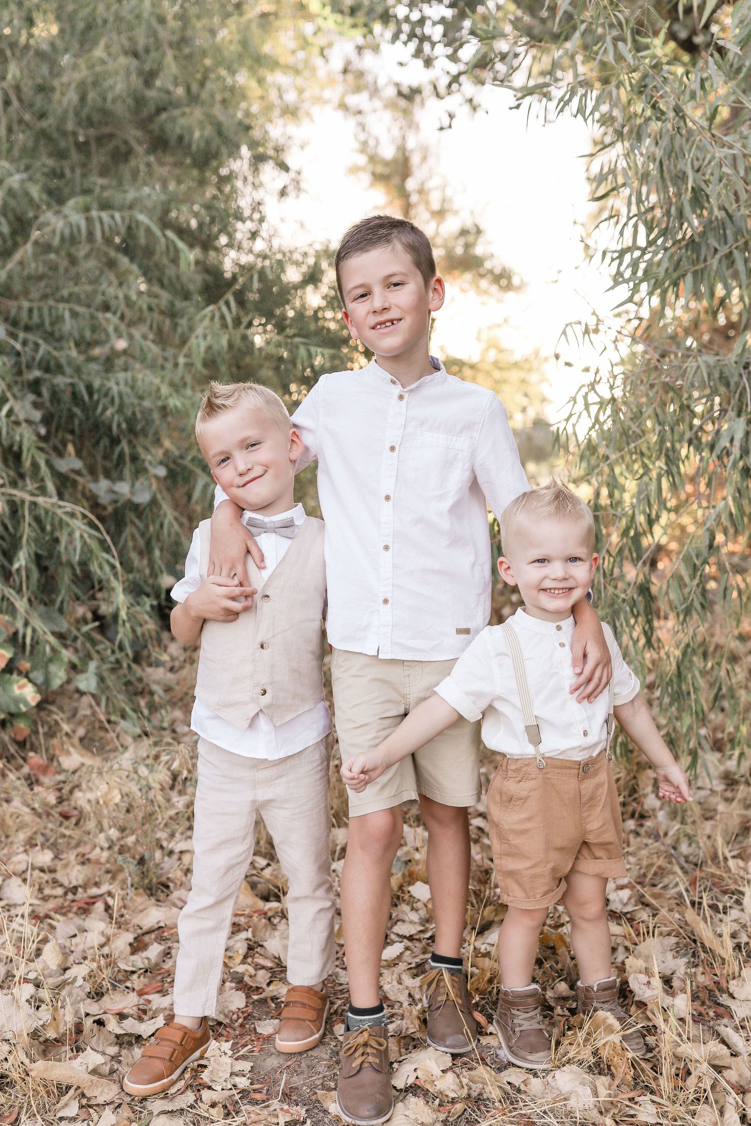 Three boys in coordinating white and tan outfits pose together outdoors among green trees and autumn leaves.