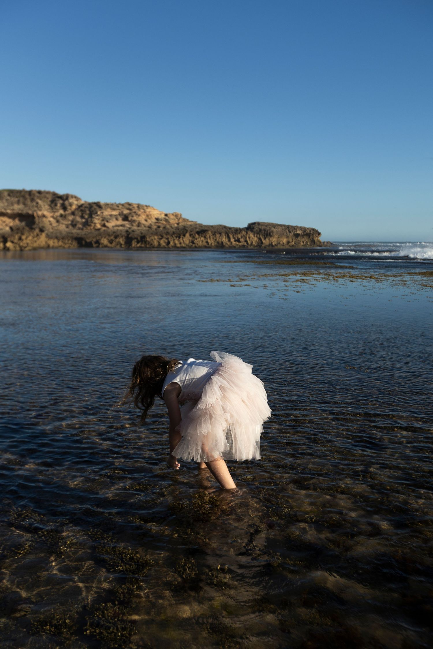 Girl in white dress wading in shallow ocean water near rocky cliffs on a clear sunny day.