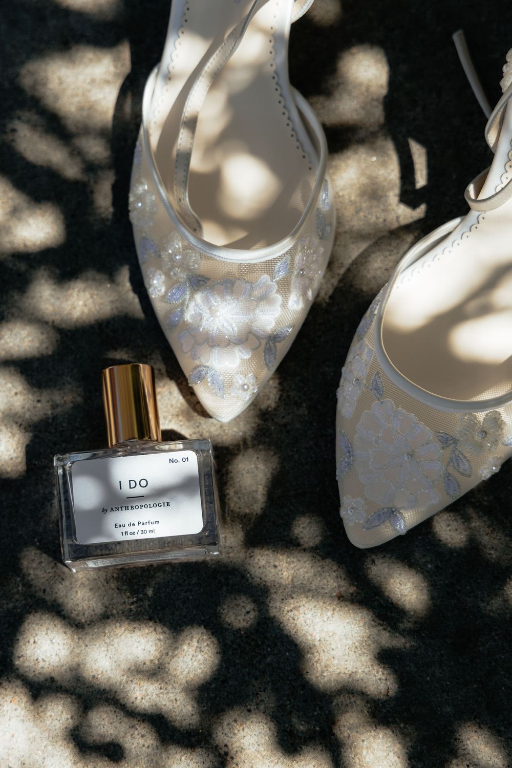 Bridal heels with floral embroidery beside an I Do perfume bottle on dappled sunlit ground.