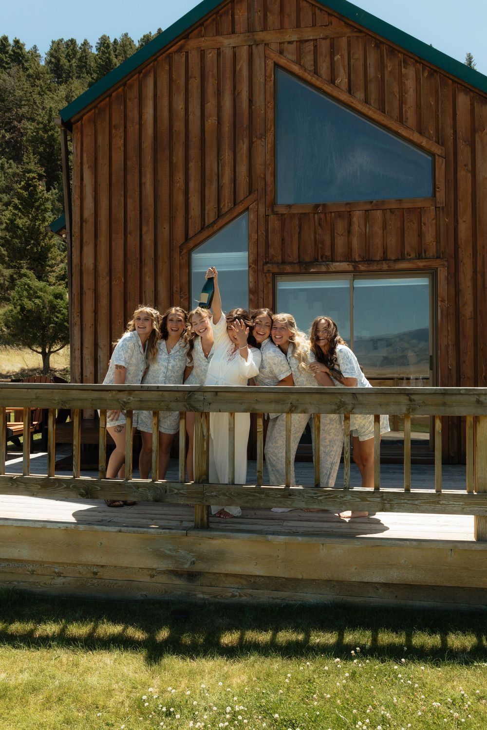 Bride and bridesmaids in matching robes posing on a wooden deck outside a rustic log cabin on a sunny day.