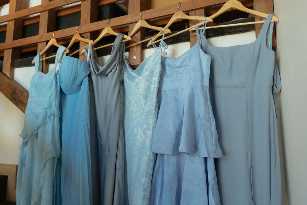 Six blue bridesmaid dresses in varying shades hanging on wooden hangers against a rustic wooden backdrop.