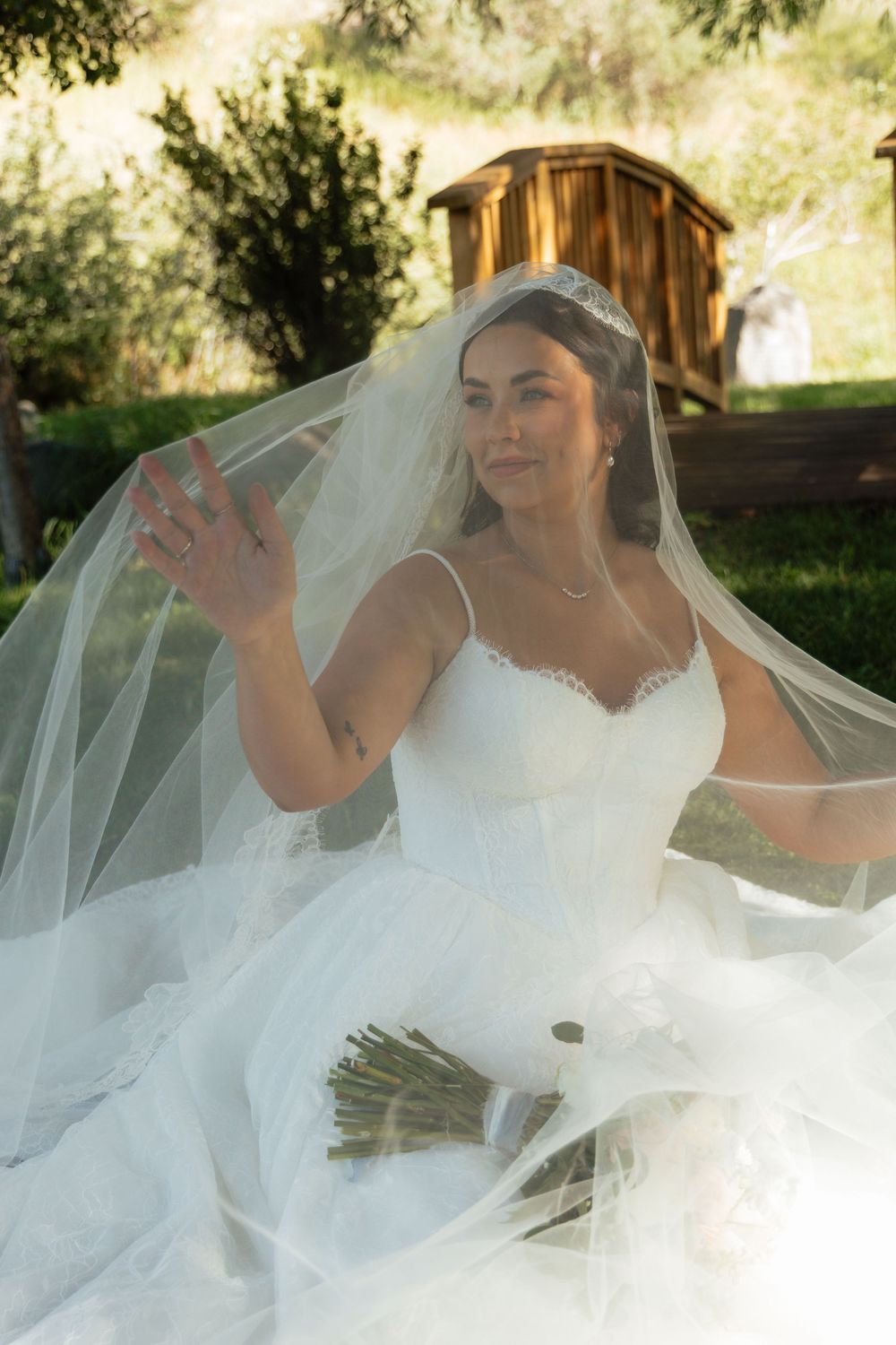 Bride in white gown and veil holds bouquet outdoors, smiling with arm raised in sunlit garden setting.
