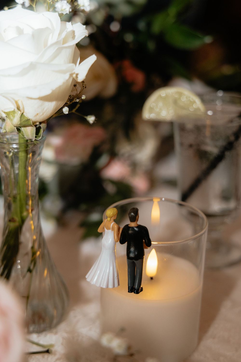 Wedding cake topper figurines of bride and groom stand before a glowing candle with white roses in background.
