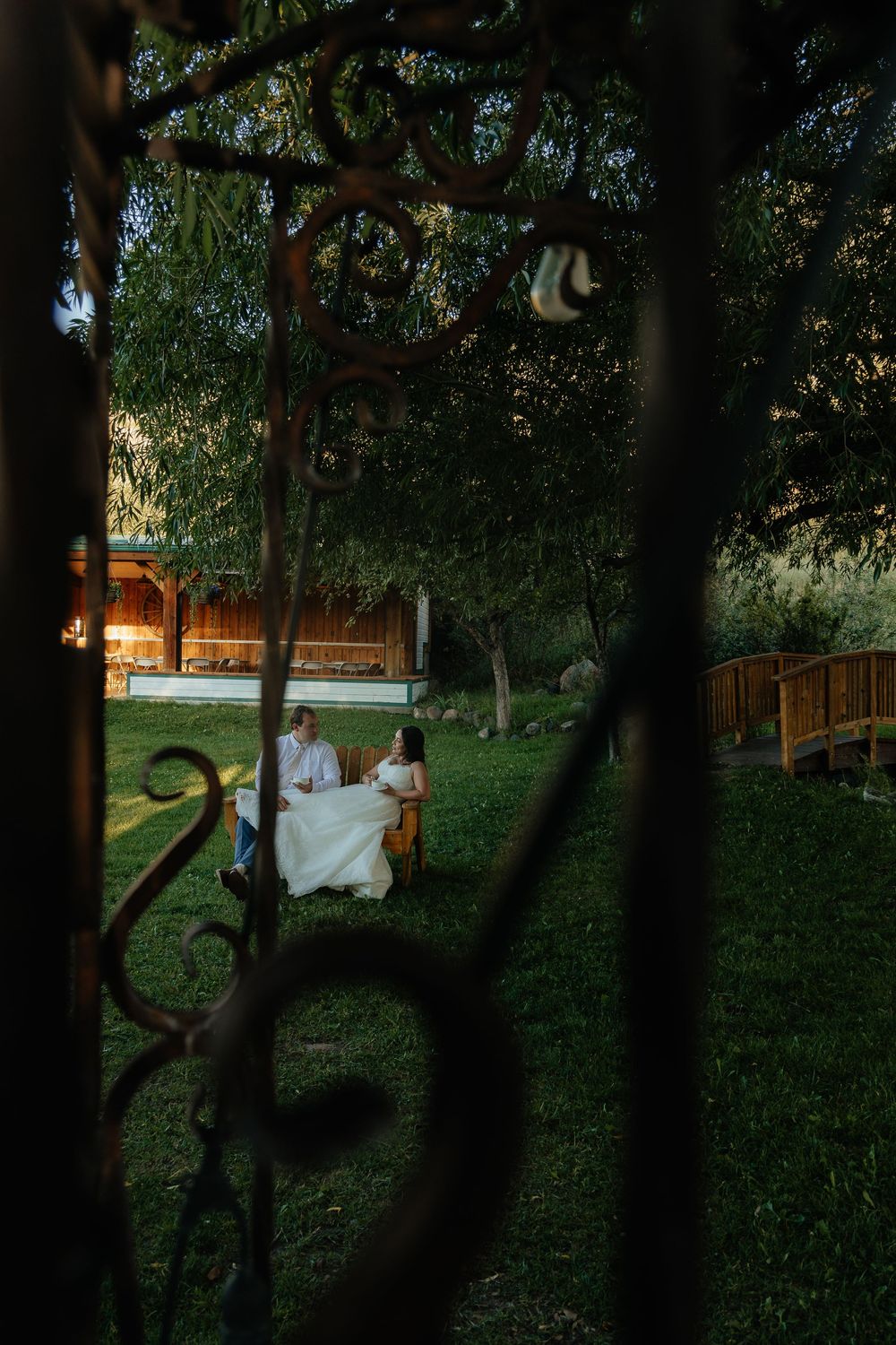 Couple in wedding attire sitting at a table in a lush garden at dusk, viewed through ornate iron gate.