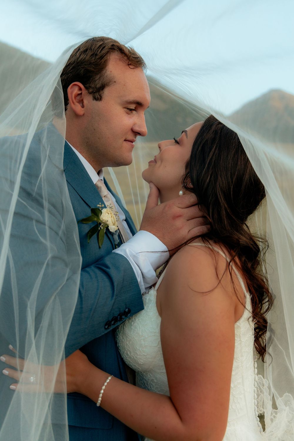 A bride and groom share a romantic moment under a flowing white veil, with the groom tenderly holding the bride's face.