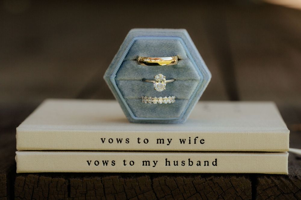 Gold wedding rings in a blue velvet hexagon box atop vow books labeled vows to my wife and vows to my husband.