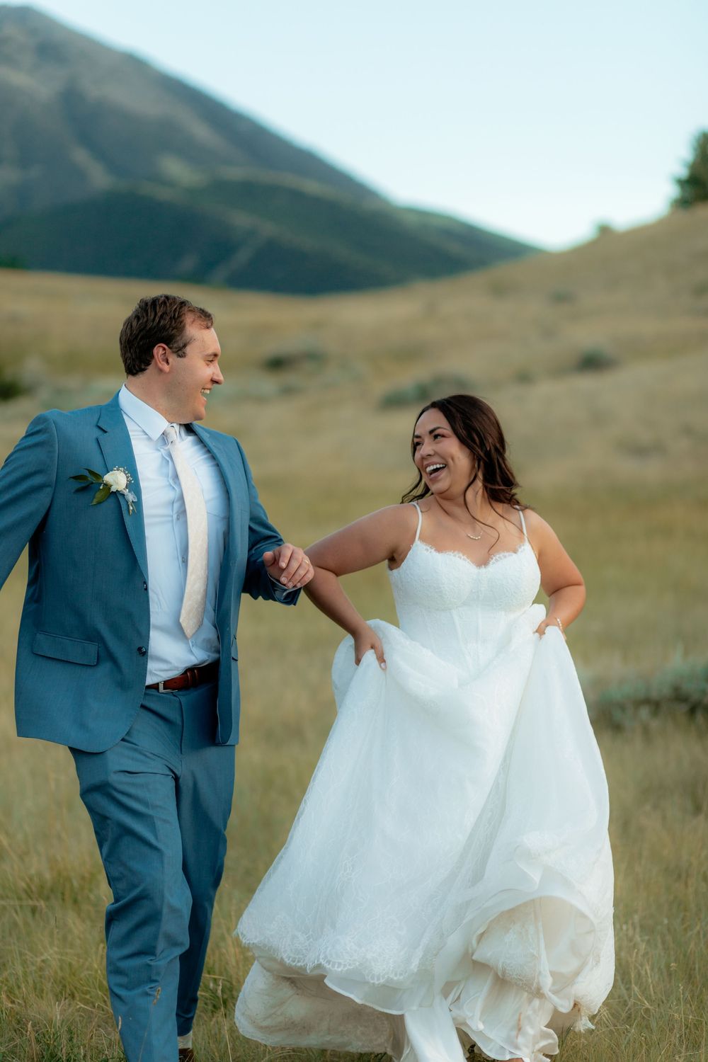 A joyful bride in a white gown and groom in a blue suit hold hands walking through a mountain meadow.