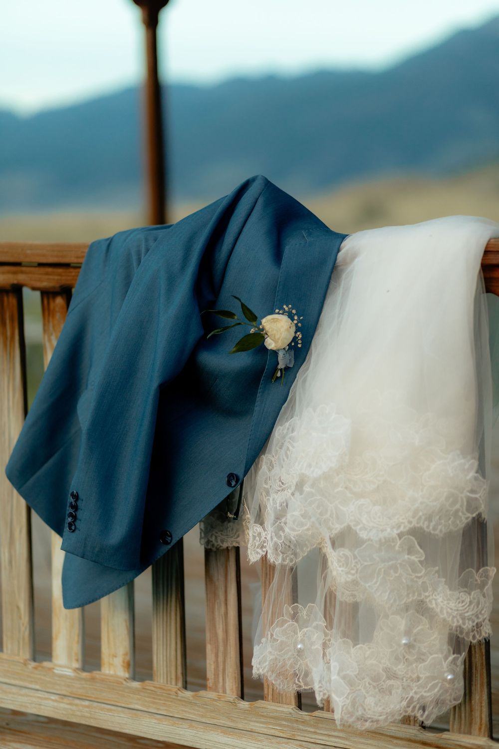 Blue groom suit with white rose boutonniere draped over wooden bench beside lace wedding veil, mountain backdrop.