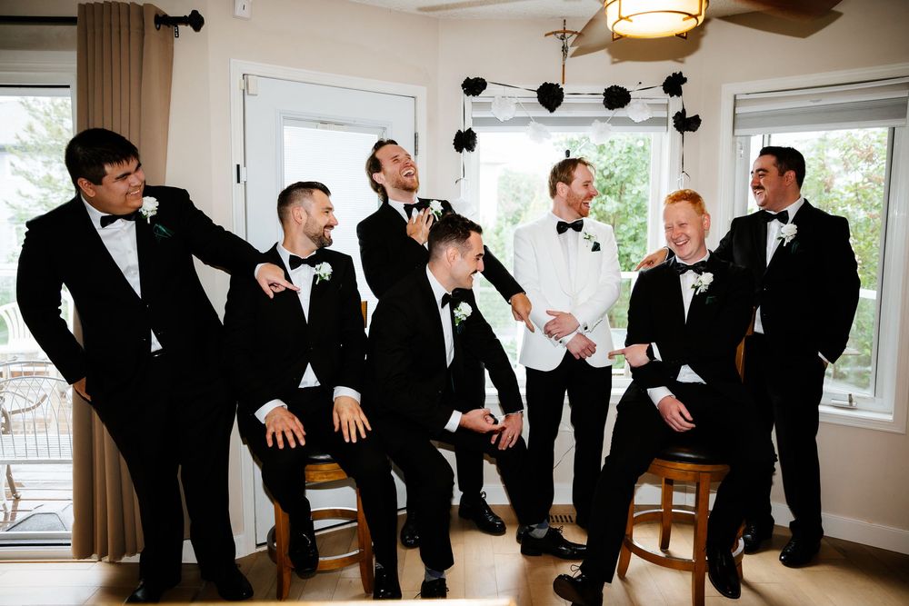 Seven groomsmen in black tuxedos with bow ties laughing and posing together in a bright room before a wedding ceremony.