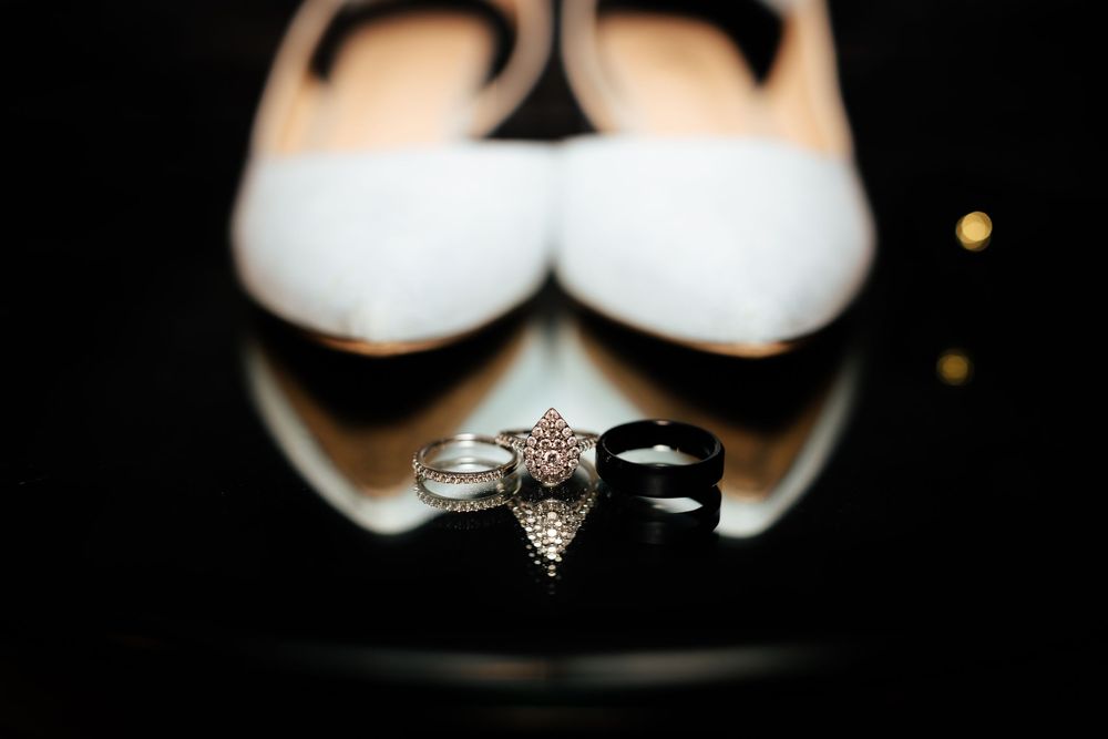 Wedding rings and bands displayed in front of white bridal heels on a reflective black surface.