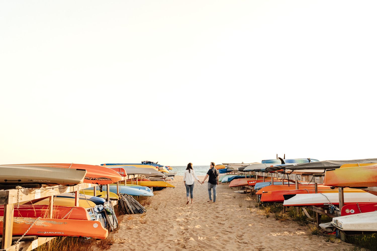 Couple holding hands walking between colorful kayaks and sailboats on a sandy beach at sunset.