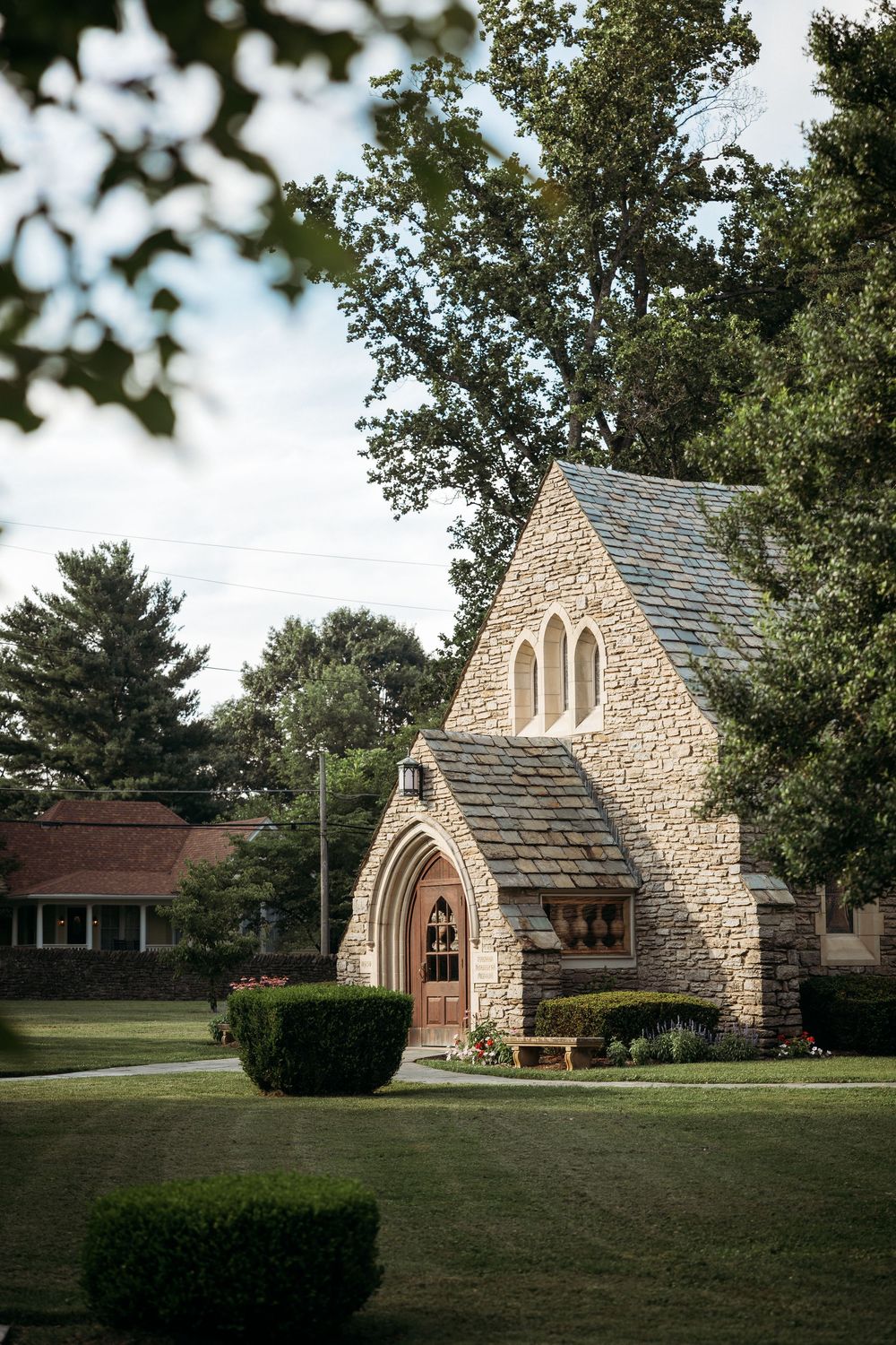 Small Gothic stone chapel with arched wooden doors, slate roof, and manicured grounds surrounded by mature trees.