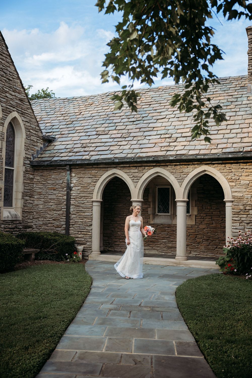 Bride in white gown holding bouquet stands on stone pathway outside elegant Gothic stone church with arched colonnade.