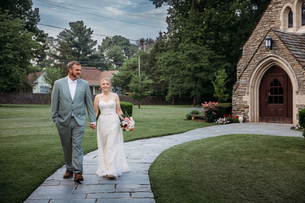 Bride and groom walking hand in hand along a stone path beside a charming chapel surrounded by lush green trees.