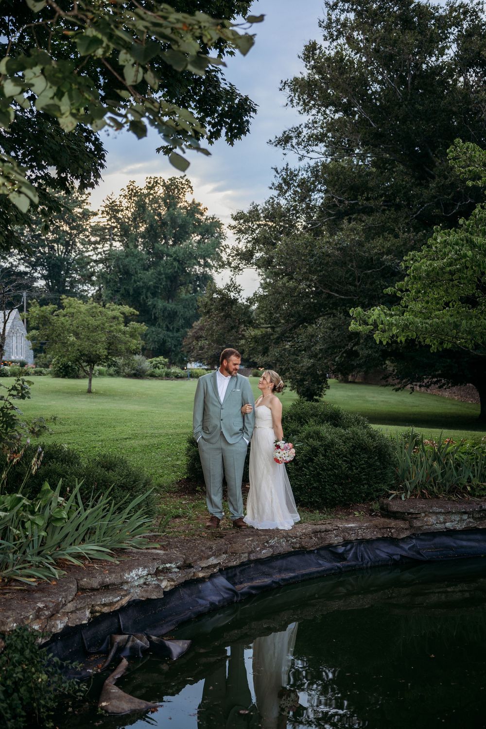 Bride and groom standing by a serene garden pond surrounded by lush green trees during their outdoor wedding.