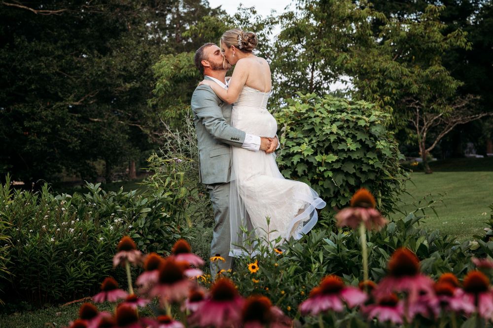 Bride and groom share a romantic kiss in a lush garden surrounded by blooming pink coneflowers on their wedding day.