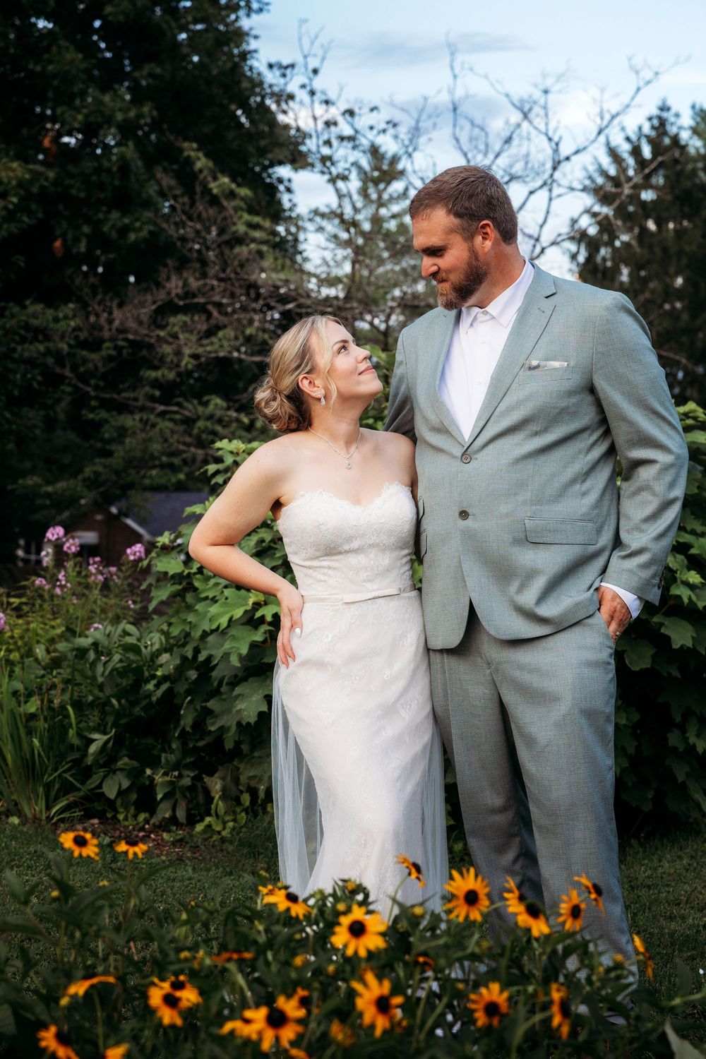 A bride and groom share a tender moment in a lush garden, surrounded by sunflowers, she in white gown, he in gray suit.