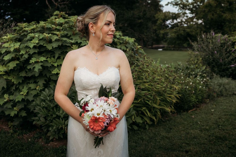 Bride in strapless white gown holding colorful bouquet of pink and white flowers, standing in lush green garden.