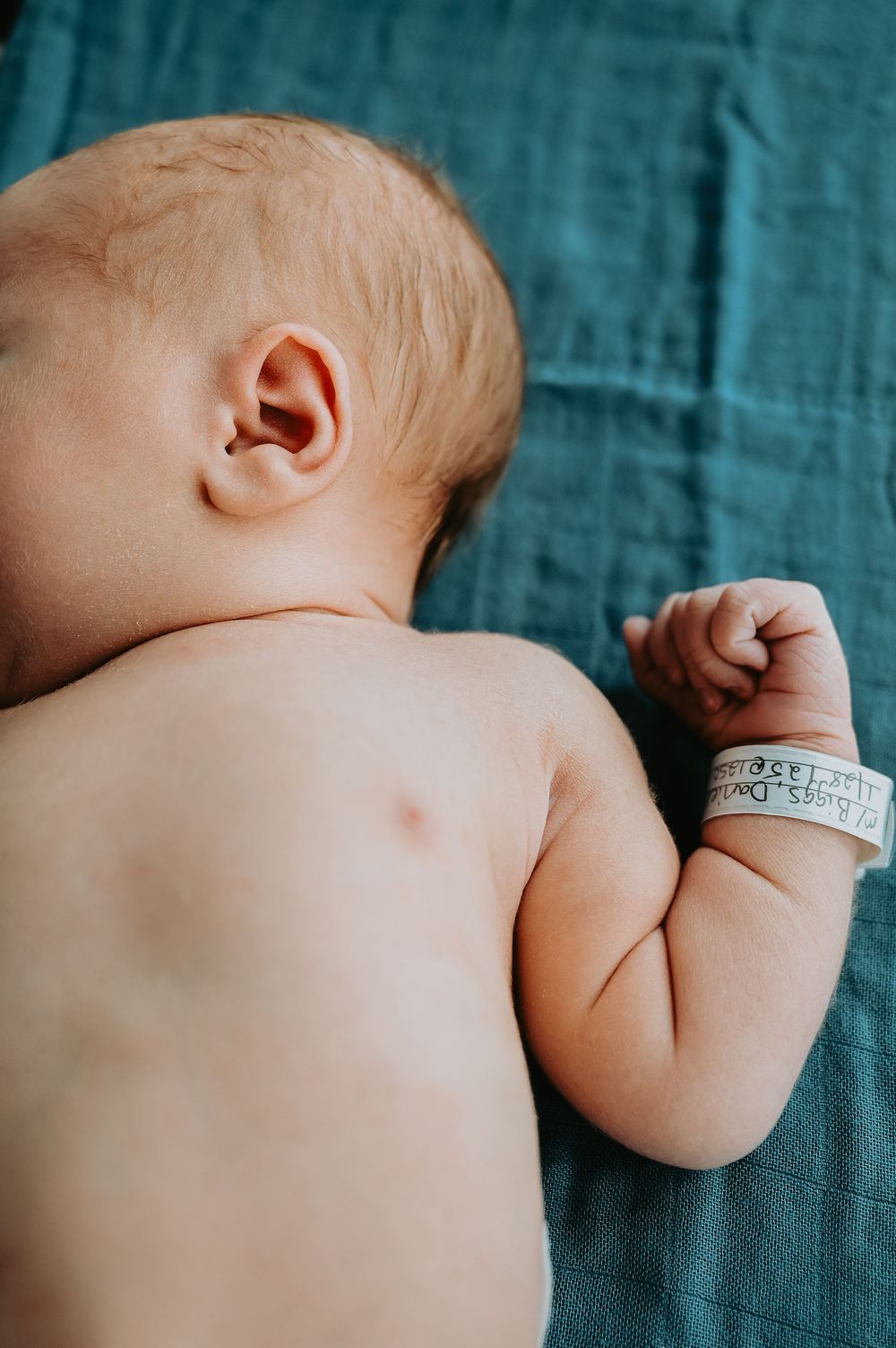 Newborn baby lying on teal blanket wearing hospital identification bracelet, viewed from behind.