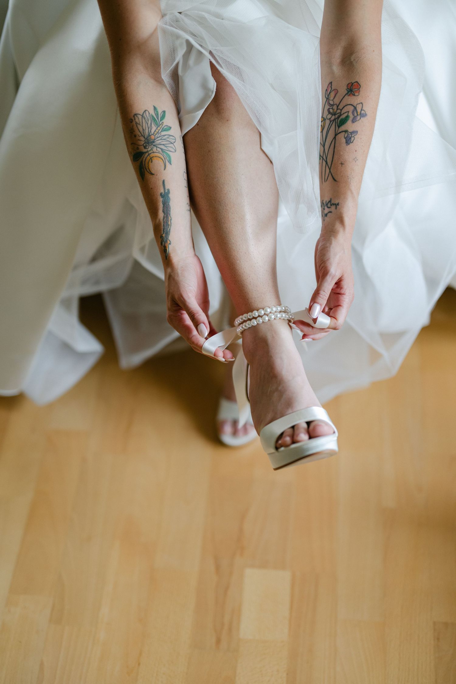 Tattooed bride in white wedding dress fastening pearl ankle strap heels on hardwood floor.