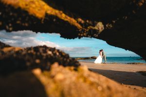 Mariée photographiée au loin sur une plage bretonne, encadrée naturellement par les rochers.