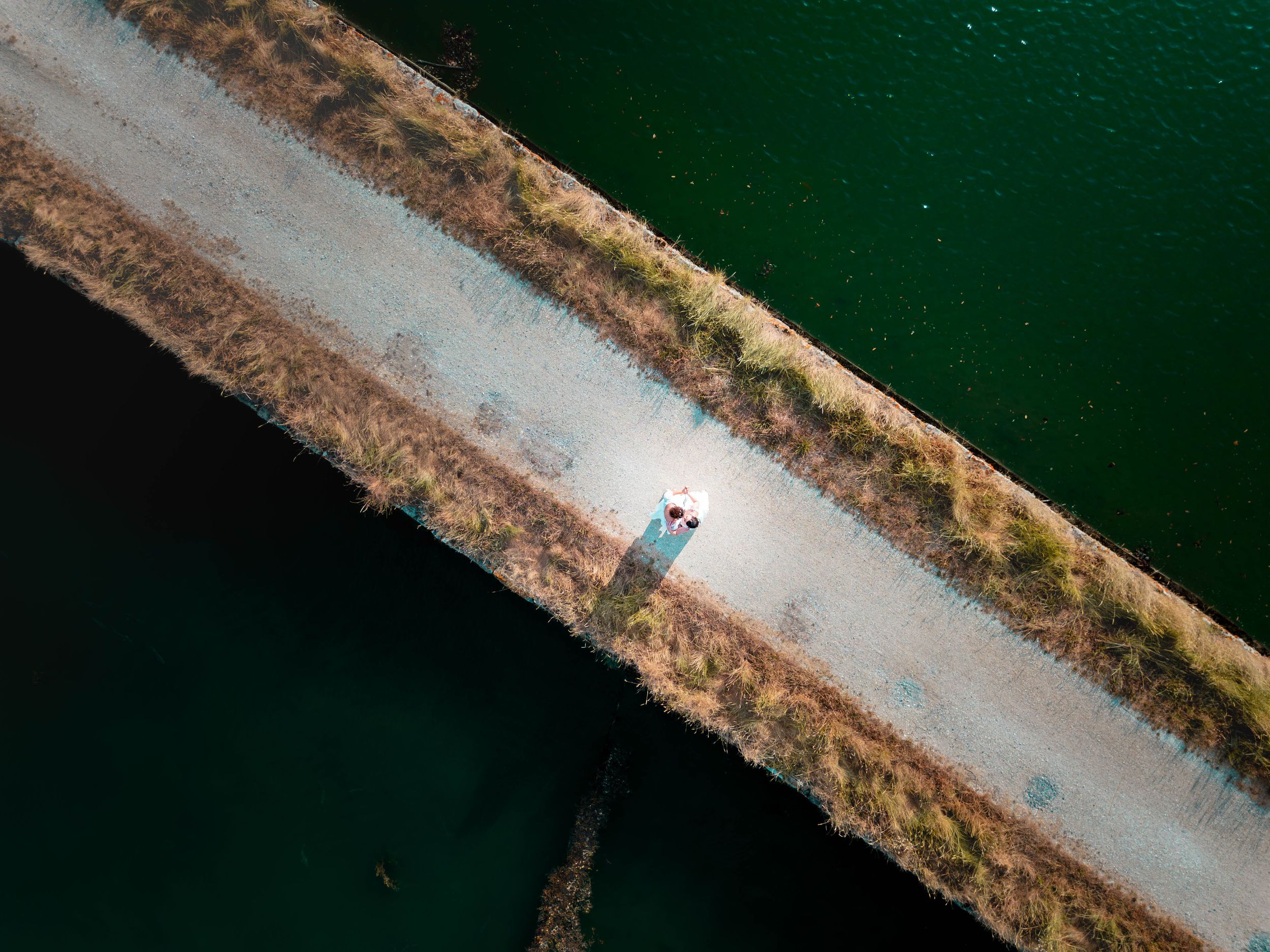 Vue aérienne par drone des mariées marchant sur un chemin entouré d’eau et de nature.