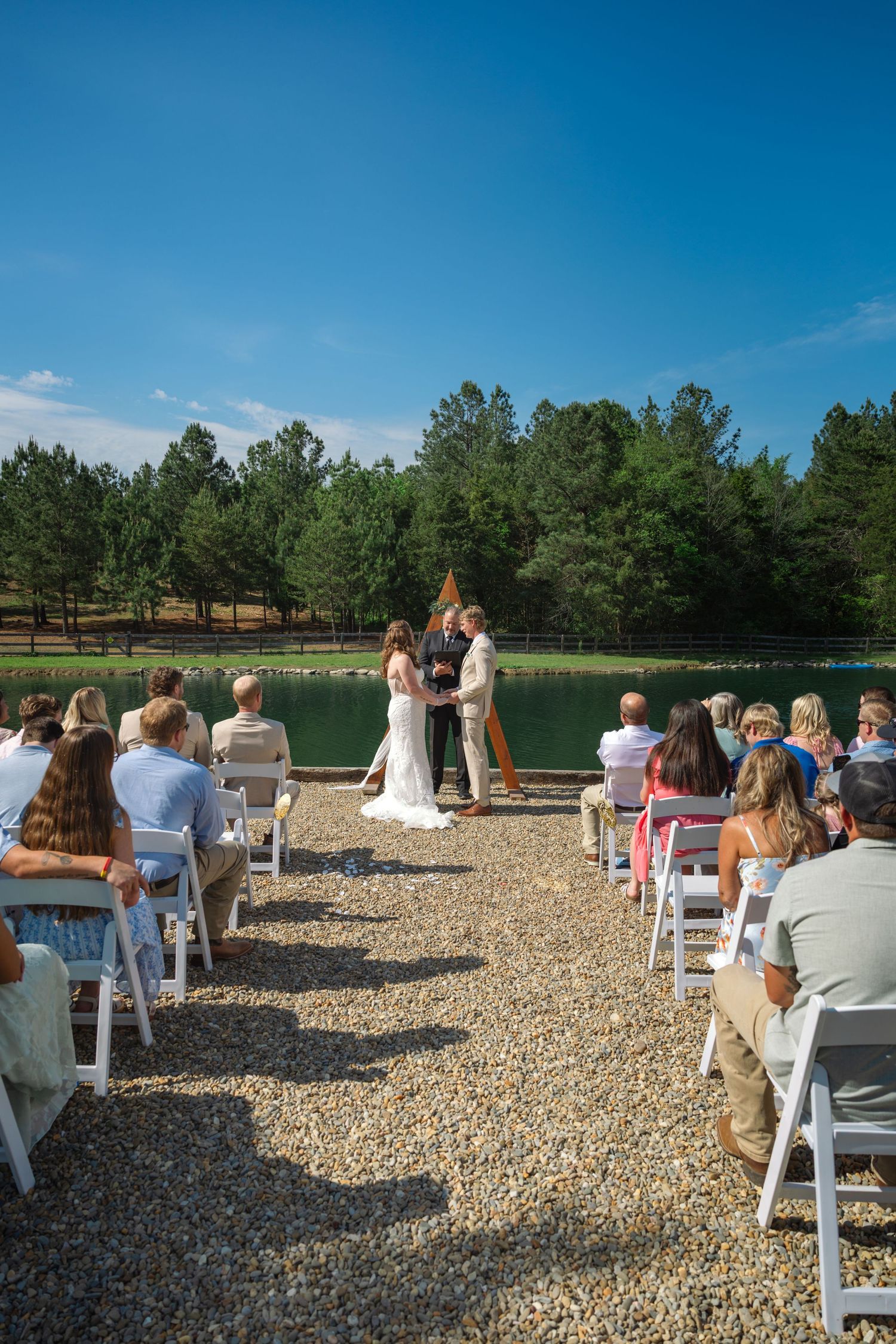 Outdoor wedding ceremony by a lake with guests seated on white chairs, surrounded by pine trees under a clear blue sky.