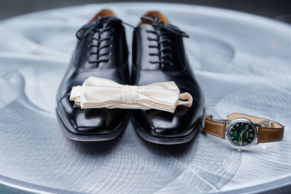 Black oxford dress shoes with cream bow tie and green dial leather watch on gray surface.