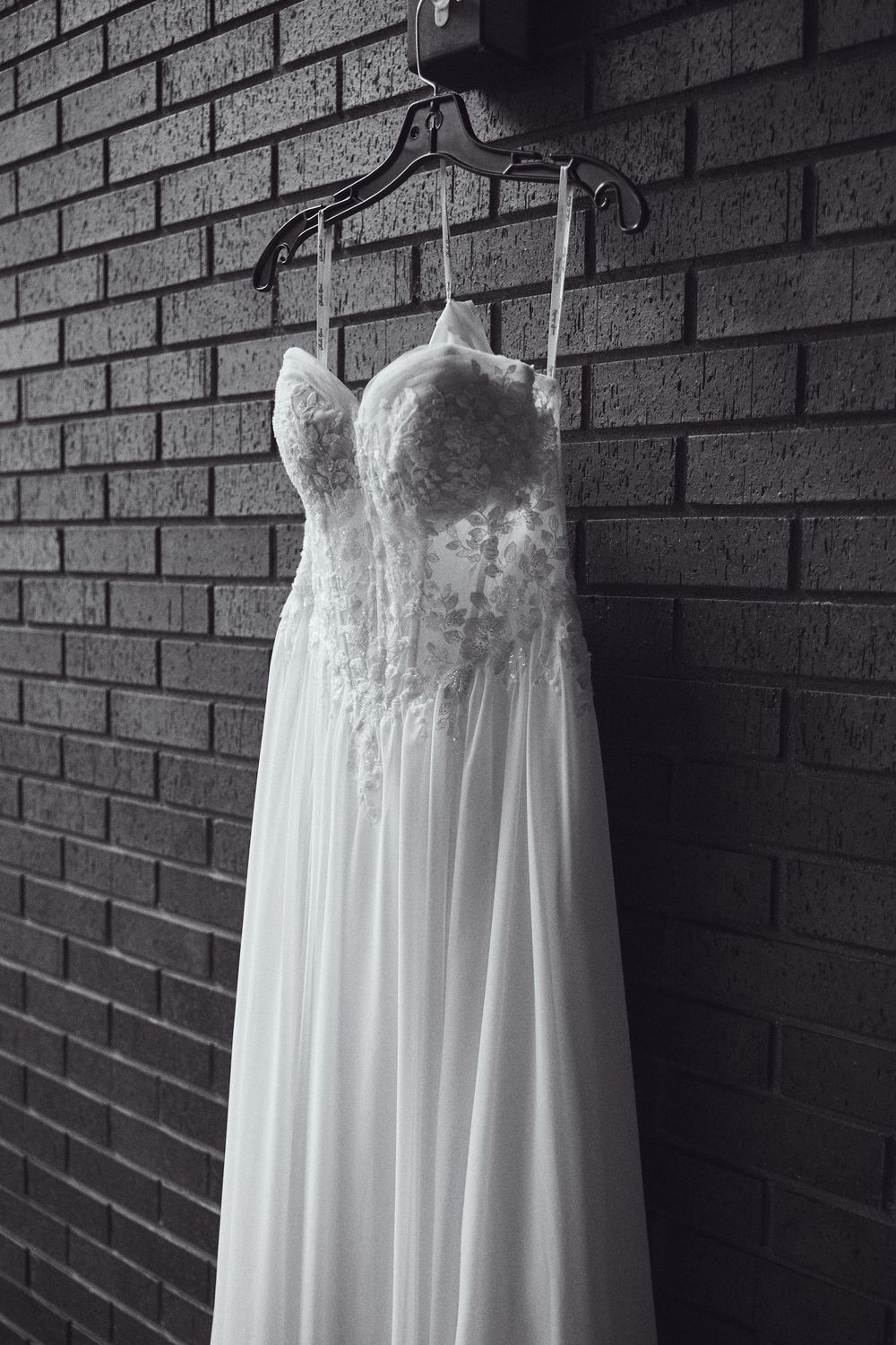 Elegant lace wedding dress hanging on a hanger against a brick wall in black and white photography.