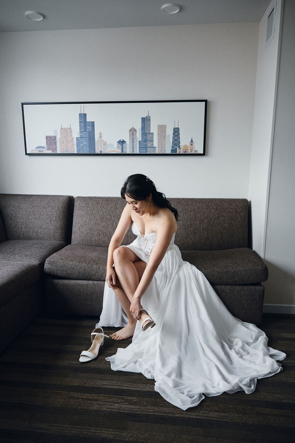 Bride in white wedding gown sitting on gray sofa adjusting her heels, with Chicago skyline artwork on wall behind her.