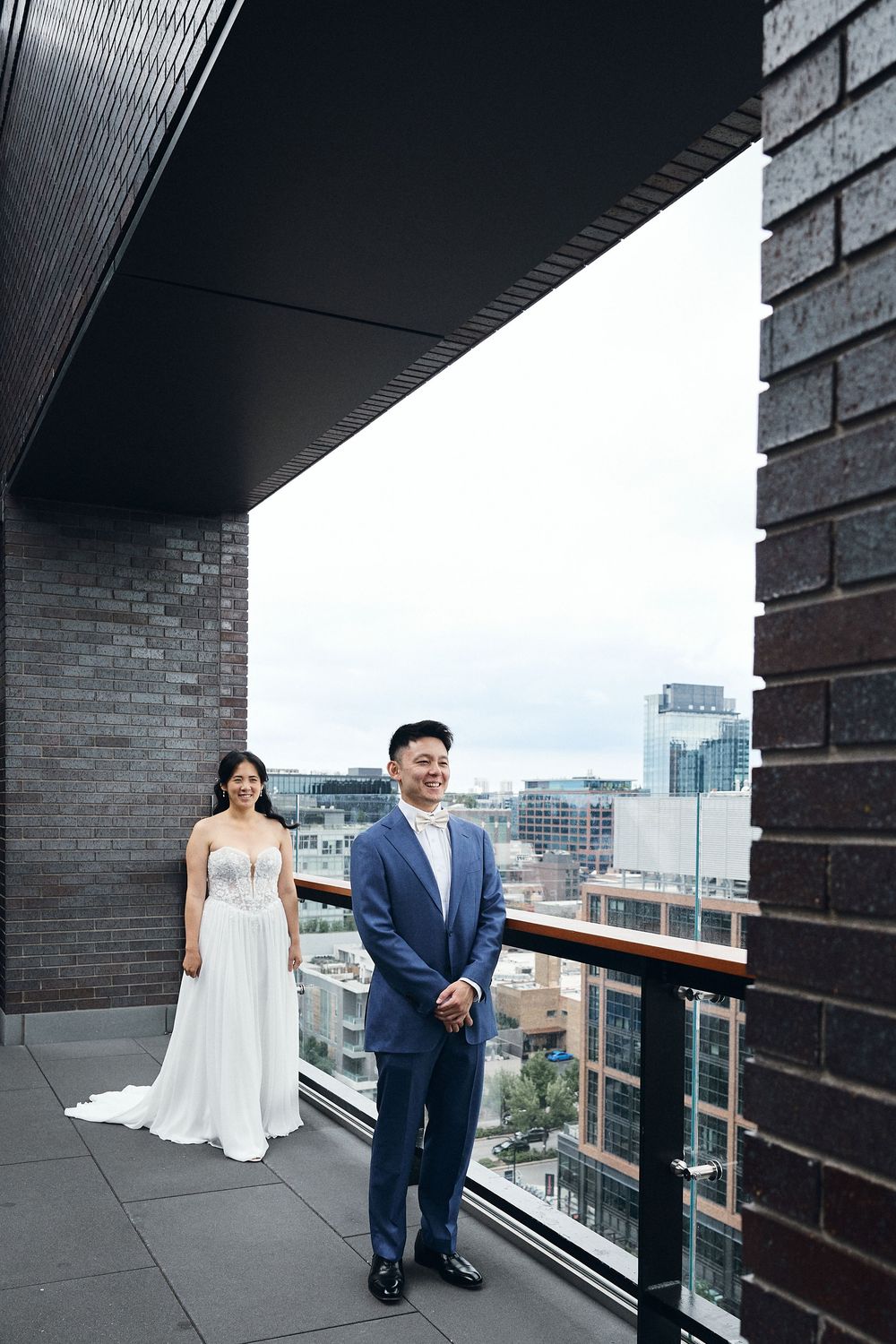 Bride in white gown and groom in blue suit pose on urban rooftop balcony with city skyline backdrop.