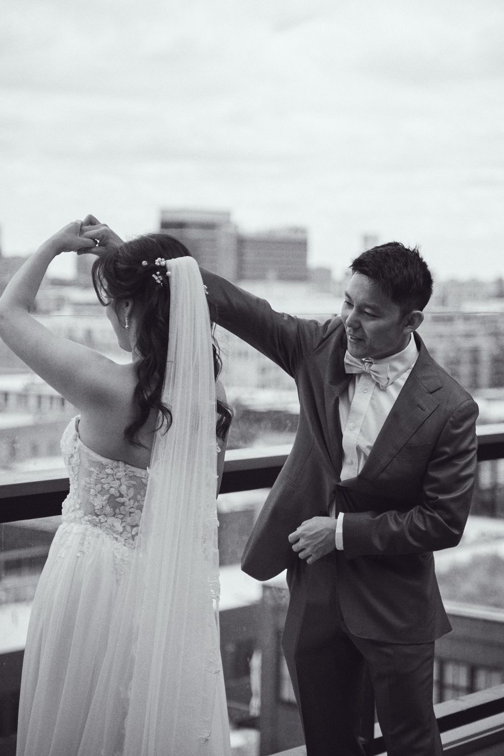 Groom adjusting bride's veil on rooftop balcony in black and white wedding photo with city skyline backdrop.