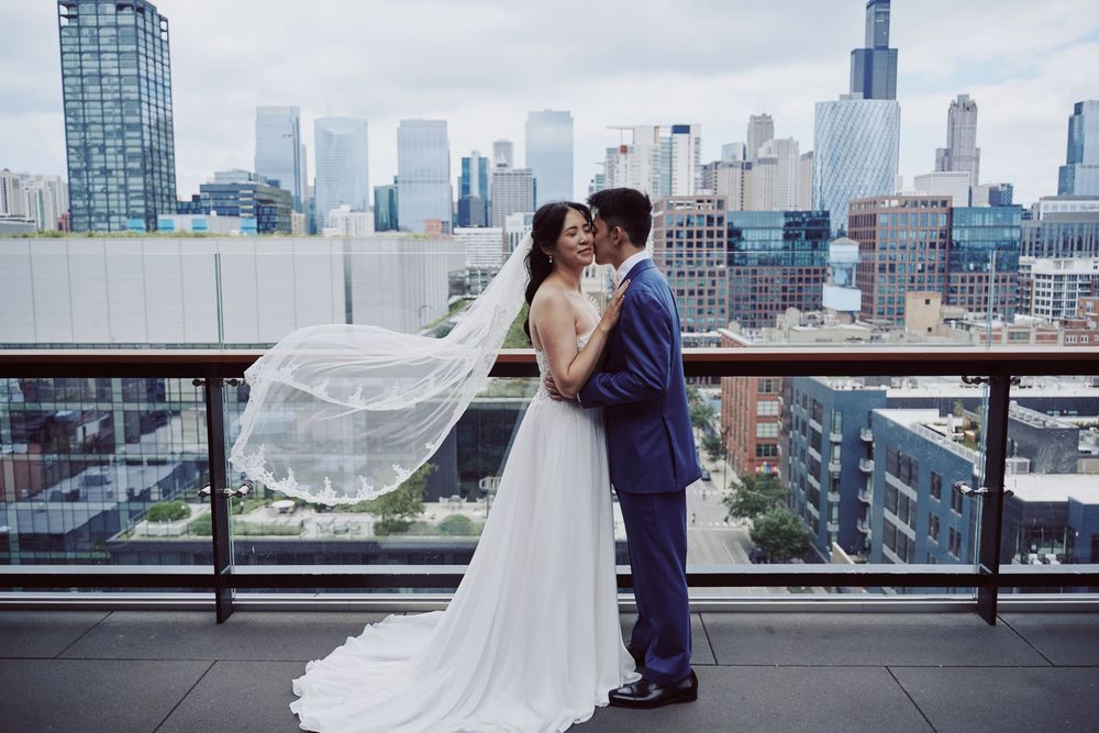 Bride and groom share a romantic kiss on a rooftop terrace with the NYC skyline backdrop, veil flowing in the wind.