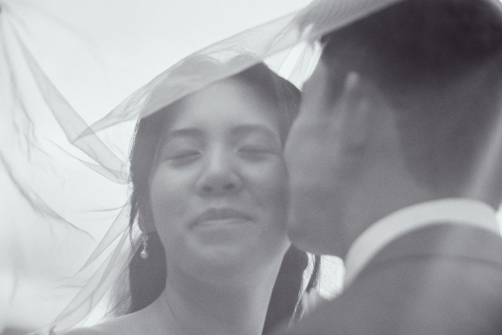 Black and white wedding photo of a bride smiling under a flowing veil, facing her groom in a romantic moment.
